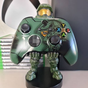 May include: A green Xbox controller with a Master Chief design. The controller is being held by a Master Chief figurine. The figurine is standing on a black base.