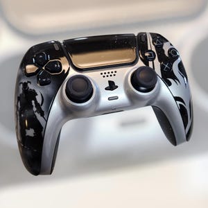 May include: A custom-designed black and silver video game controller. The controller features a silver center section with black joysticks and buttons. The sides are decorated with black and white graphics. The PlayStation logo is visible.