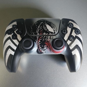 Puede incluir: Un mando de PlayStation 5 en blanco y negro con un diseño de Venom. El mando tiene una base negra con detalles blancos y una ilustración de Venom en el centro. Los botones y joysticks son negros.