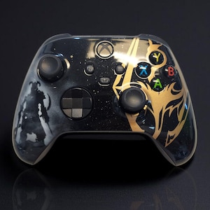 May include: Black Xbox controller with a custom design. The controller has a gold and black design with a warrior silhouette on the left and a gold graphic on the right. The buttons are multi-coloured.