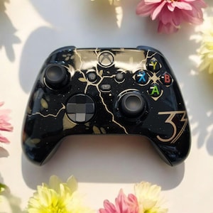 Może przedstawiać: Czarny kontroler Xbox ze złotym i białym marmurowym wzorem. Kontroler ma złoty numer 33 po prawej stronie. Przyciski są w kolorach zielonym, niebieskim, żółtym i czerwonym. Kontroler jest otoczony różowymi i żółtymi kwiatami.