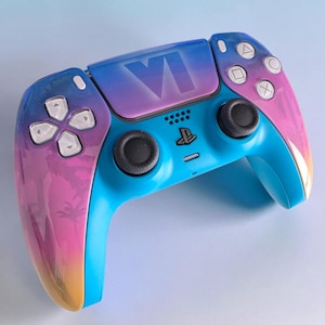 May include: A wireless video game controller featuring a vibrant gradient finish that transitions from blue to pink and orange. The central touchpad displays the Roman numeral VI. It includes two black analog sticks, a directional pad, and white buttons.