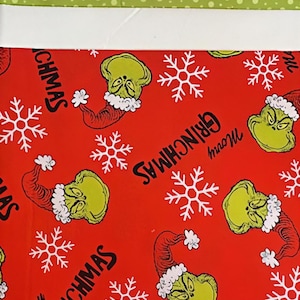 Puede incluir: Tela roja con un patrón repetido de la cara del Grinch con gorro de Papá Noel, copos de nieve y las palabras "GRINCHMAS" y "Merry". Borde superior con lunares verdes y blancos.