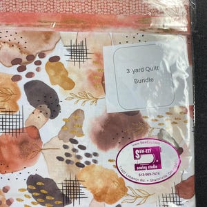 May include: A 3-yard quilt bundle in a clear plastic package. The fabric features an abstract design with watercolor-style shapes in shades of brown, tan, and gold, with black grid and leaf accents. The label reads "3 yard Quilt Bundle."