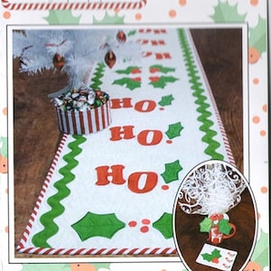 May include: A white Christmas table runner with red and green holly leaf and "HO HO HO" designs. The runner is bordered with a red and white striped pattern and green zig-zag trim. The image also shows a Christmas tree and a small gift box.