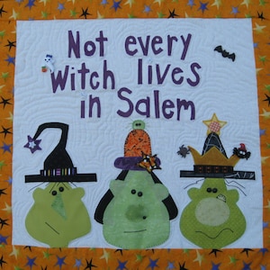 Pode incluir: Uma tapeçaria quadrada de tecido com uma borda laranja decorada com estrelas coloridas. O centro branco exibe o texto roxo "Not every witch lives in Salem" acima de três rostos de desenhos animados verdes com chapéus decorativos e adornos.