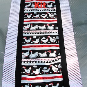 May include: Table runner kit featuring a black fabric with white chickens and red accents. The kit includes two white fabric strips with red polka dots.
