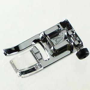 May include: A polished, chrome sewing machine presser foot with a clear plastic sole. The foot features a curved lever and a black knob. The letter "A" is visible on the metal. This sewing machine accessory is designed for various sewing projects.