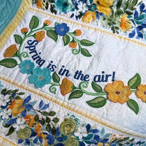May include: White quilt with floral embroidery and the phrase "Spring is in the air!" in blue script. The quilt features embroidered flowers in shades of blue, yellow, and orange, with green leaves. The edges are trimmed with yellow.