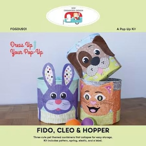 May include: Three pet-themed pop-up containers: a dog, a cat, and a bunny. The containers are cylindrical and feature colorful fabric designs. The dog container is brown and yellow, the cat is orange, and the bunny is purple. The kit includes a pattern, spring, elastic, and a label.