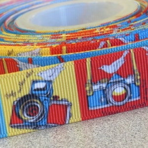 May include: A colorful ribbon with a repeating pattern of cameras and white doves. The ribbon is blue, yellow, red, and white. The cameras are red, blue, and black. The doves are white with black eyes. The ribbon has the text "Smile for the shot" printed on it.