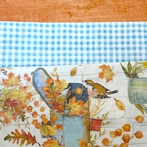 Puede incluir: Una servilleta de papel decorativa con un buzón azul y dos pájaros, rodeada de hojas de otoño y bayas. El fondo tiene un patrón de vetas de madera blanca. Se puede ver un patrón de cuadros azul claro y blanco.