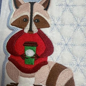 May include: Embroidered patch featuring a raccoon wearing a red sweater and holding a green coffee cup. The raccoon has brown and tan fur with a black mask. The background is white with blue snowflake patterns.