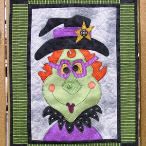 May include: A green witch with a purple dress, black hat, and orange hair. She is wearing glasses and has a star on her hat. The witch is framed in a green and black striped border.