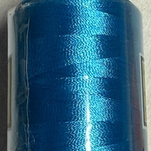 Puede incluir: Un carrete de hilo de bordar turquesa vibrante. El hilo está enrollado firmemente en un carrete cilíndrico, envuelto en plástico transparente. La parte superior e inferior del carrete son blancas. El hilo tiene una calidad brillante y reflectante, para coser y manualidades.