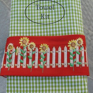 May include: A green and white gingham towel with a red border featuring a white picket fence and sunflowers. The towel has the text "Towel Kit" printed on it.