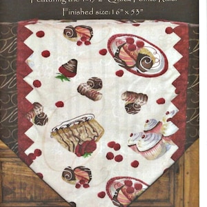 Puede incluir: Un camino de mesa blanco con un borde rojo y un patrón de postres de chocolate y frambuesas rojas. El texto "Prairie's On-Point Table Runner" y "Featuring the 1-1/2" Quick Points Ruler" está impreso en la parte superior de la imagen. El texto "Finished size: 16" x 53" está impreso debajo del título.