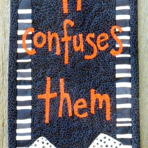May include: A black and white striped fabric wall hanging with the text "Smile - it confuses them" in orange embroidery. A white dog face with black polka dots and blue eyes is at the bottom of the hanging.