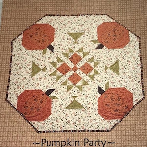 May include: A quilted pumpkin-themed table runner with four orange pumpkins and green accents. The center features a geometric design with orange and green shapes. The text "Pumpkin Party" is at the bottom, with "This & That" at the top.