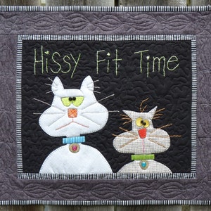 May include: A quilted wall hanging featuring two cartoon cats with the text "Hissy Fit Time". The cats are white and tan with green and yellow eyes, wearing collars with decorative buttons. The background is black with a decorative border.