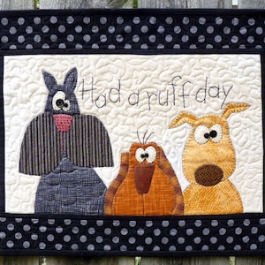 May include: A handmade quilted wall hanging featuring three whimsical dog-like creatures. The phrase "Had a ruff day" is embroidered above the characters. The border is black with white polka dots, and the background is quilted.
