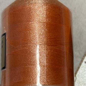 Può includere: Una bobina di filo da ricamo color arancione. Il filo è avvolto strettamente attorno a un'anima di plastica bianca e racchiuso in un involucro di plastica trasparente. Il filo ha una qualità lucida e riflettente.
