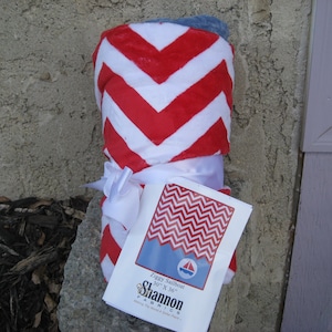 Pode incluir: Um cobertor de chevron vermelho e branco dobrado e amarrado com uma fita branca. O cobertor é etiquetado "Ziggy Sailboat 30" x 36" Shannon Fabrics"