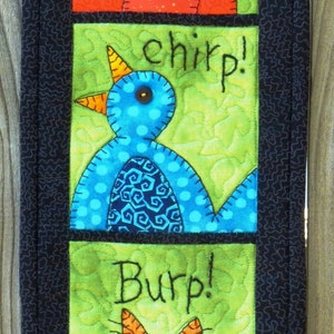 May include: A quilted wall hanging with three panels featuring a red bird, a blue bird, and an orange cat. The panels are bordered in black fabric and each panel has a word embroidered on it: "Chirp!" "chirp!" and "Burp!"