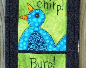 Puede incluir: Un tapiz de pared acolchado con tres paneles que representan un pájaro rojo, un pájaro azul y un gato naranja. Los paneles están bordeados con tela negra y cada panel tiene una palabra bordada en él: "Chirp!" "chirp!" y "Burp!"