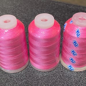 Op de afbeelding: Drie spoelen roze garen met witte plastic bovenkanten. Het garen is om de spoelen gewikkeld en de spoelen hebben blauwe etiketten met de tekst "YLI" erop gedrukt.
