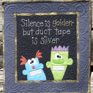 May include: Quilted wall hanging with the text "Silence is golden - but duct tape is silver." Features two whimsical monster figures in blue and green. The piece has a black background with a gold and blue border.