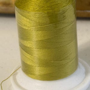 May include: A spool of olive green sewing thread wound onto a white plastic cylinder. The thread is tightly coiled around the centre, showing a smooth, consistent texture and a vibrant, earthy green colour. A single loose strand extends from the base.