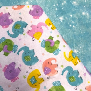 May include: A white fabric with a repeating pattern of colorful cartoon elephants in purple, pink, yellow, green, and blue. The elephants have polka dot accents and are scattered among small stars. A light blue fabric with white star-like shapes is in the background.