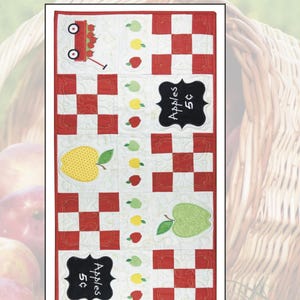 May include: A quilted table runner with an apple theme. The runner features red and white checkered squares, apple appliqués in various colors, and the text "Apples 5¢" on a chalkboard-style design. The top banner reads "Apple-a-Day Table Runner".