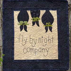 May include: A beige quilted wall hanging with a black border and three black bats with green eyes and hats. The text "fly by night company" is embroidered in black on the beige fabric.