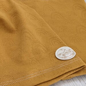 Puede incluir: Tela amarilla mostaza con un sutil patr&oacute;n en relieve de peque&ntilde;os corazones. La tela tiene una costura blanca cosida y una peque&ntilde;a etiqueta blanca con una ilustraci&oacute;n en blanco y negro de una mariquita.