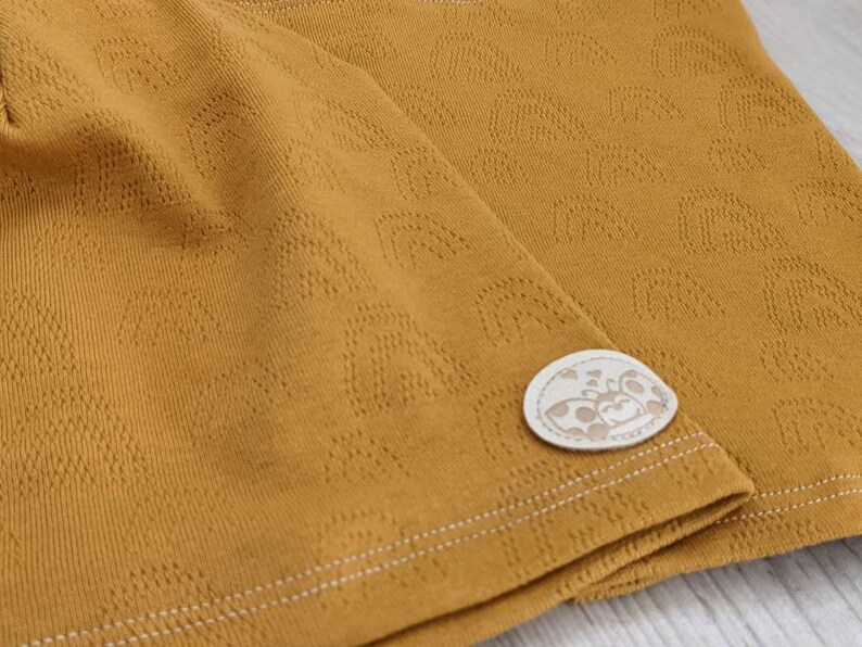 Puede incluir: Tela amarilla mostaza con un sutil patr&oacute;n en relieve de peque&ntilde;os corazones. La tela tiene una costura blanca cosida y una peque&ntilde;a etiqueta blanca con una ilustraci&oacute;n en blanco y negro de una mariquita.