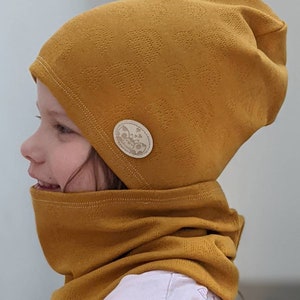Puede incluir: Un gorro de punto amarillo mostaza con un cuello de punto a juego. El gorro tiene un peque&ntilde;o bot&oacute;n redondo de color crema con un dise&ntilde;o de mariposa en el lateral.