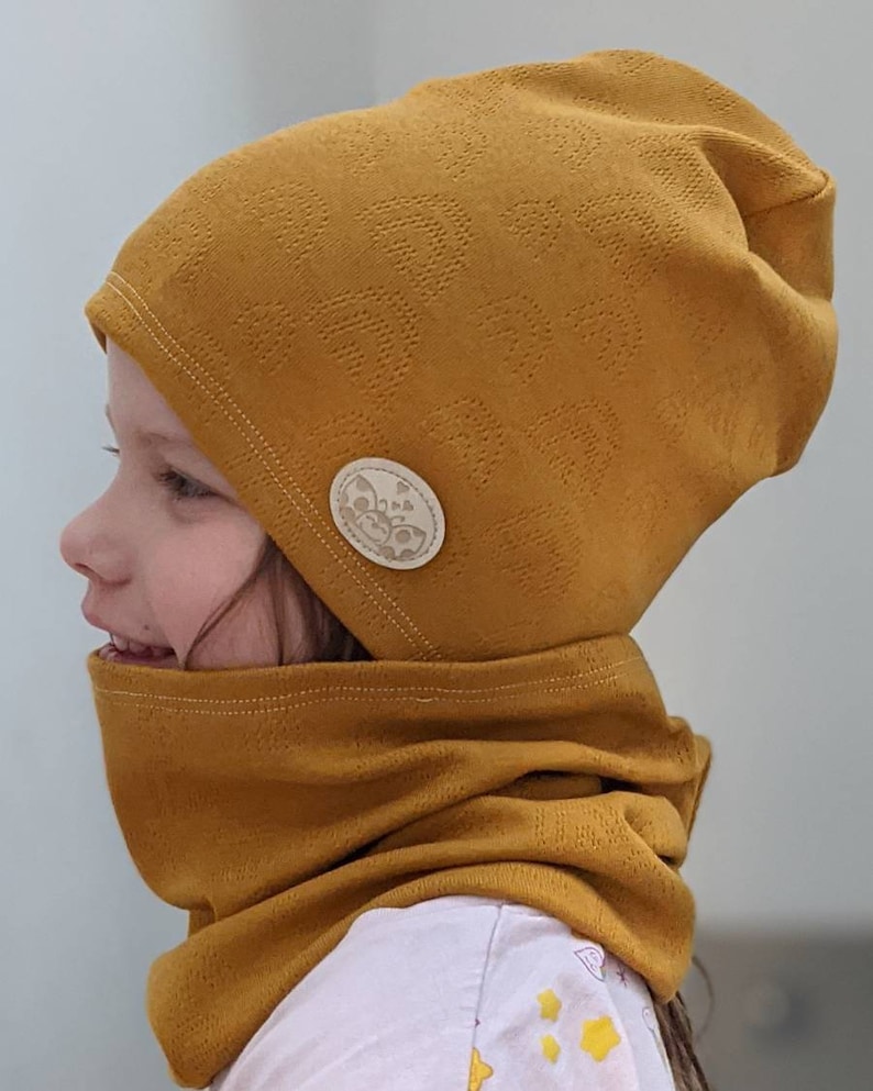 Puede incluir: Un gorro de punto amarillo mostaza con un cuello de punto a juego. El gorro tiene un peque&ntilde;o bot&oacute;n redondo de color crema con un dise&ntilde;o de mariposa en el lateral.