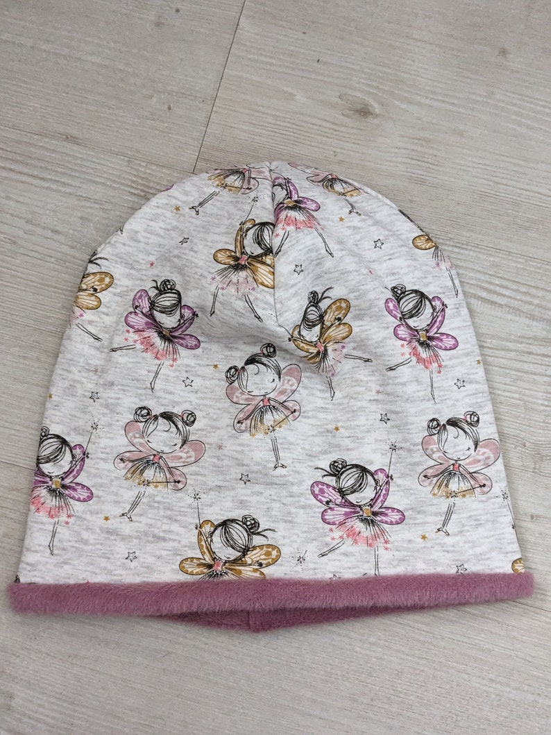 Puede incluir: Un gorro gris con forro de borreguito rosa. El gorro tiene un patr&oacute;n de hadas con alas y estrellas.