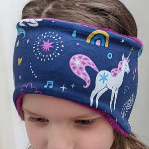 May include: A navy blue headband with a unicorn and rainbow pattern. The headband has a pink fleece lining and features a white unicorn with a pink mane and tail. Other design elements include stars, musical notes, and a rainbow.