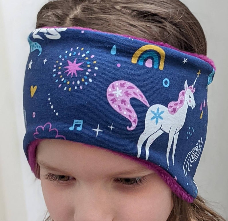 May include: A navy blue headband with a unicorn and rainbow pattern. The headband has a pink fleece lining and features a white unicorn with a pink mane and tail. Other design elements include stars, musical notes, and a rainbow.