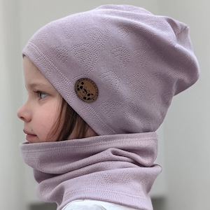 Puede incluir: Un gorro de punto morado con una peque&ntilde;a etiqueta marr&oacute;n que presenta un dise&ntilde;o gr&aacute;fico. El gorro lo lleva puesto un ni&ntilde;o y se combina con un cuello de punto morado a juego.
