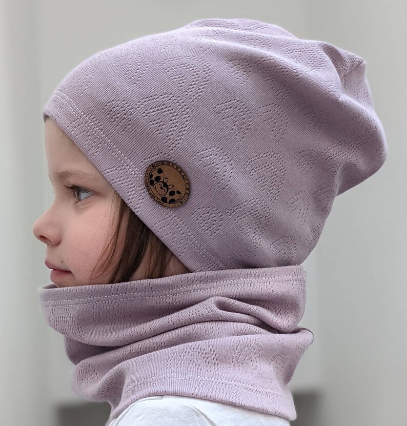Puede incluir: Un gorro de punto morado con una peque&ntilde;a etiqueta marr&oacute;n que presenta un dise&ntilde;o gr&aacute;fico. El gorro lo lleva puesto un ni&ntilde;o y se combina con un cuello de punto morado a juego.