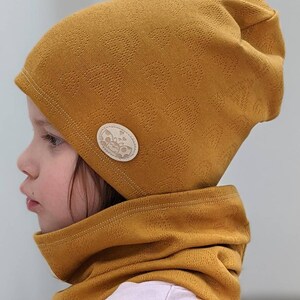Puede incluir: Un gorro de punto amarillo mostaza y un cuello de punto a juego con un peque&ntilde;o bot&oacute;n redondo beige con un dise&ntilde;o de mariposa.