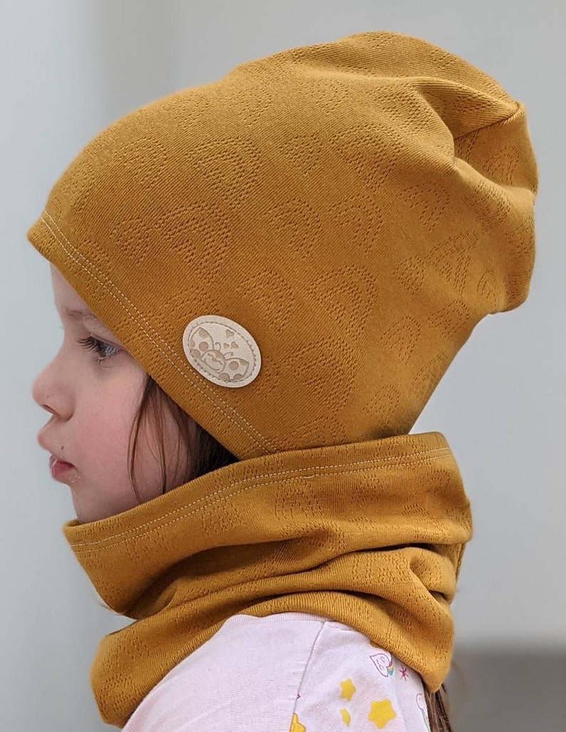 Puede incluir: Un gorro de punto amarillo mostaza y un cuello de punto a juego con un peque&ntilde;o bot&oacute;n redondo beige con un dise&ntilde;o de mariposa.