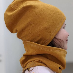 Puede incluir: Un conjunto de gorro y cuello de color mostaza. El gorro est&aacute; doblado hacia abajo y el cuello est&aacute; subido alrededor del cuello.