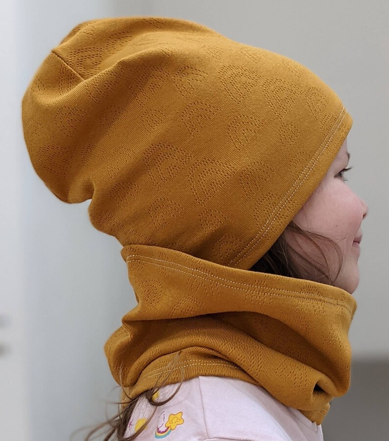 Puede incluir: Un conjunto de gorro y cuello de color mostaza. El gorro est&aacute; doblado hacia abajo y el cuello est&aacute; subido alrededor del cuello.