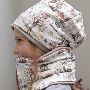 May include: A white beanie and neck warmer set with a woodland animal print. The fabric features a light brown background with illustrations of deer, foxes, raccoons, and trees.