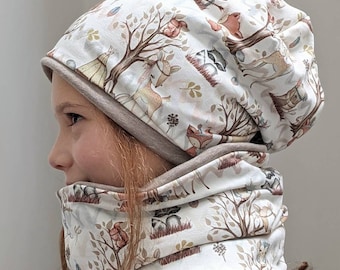 Puede incluir: Un conjunto de gorro y cuello blanco con estampado de animales del bosque. La tela presenta un fondo marrón claro con ilustraciones de ciervos, zorros, mapaches y árboles.